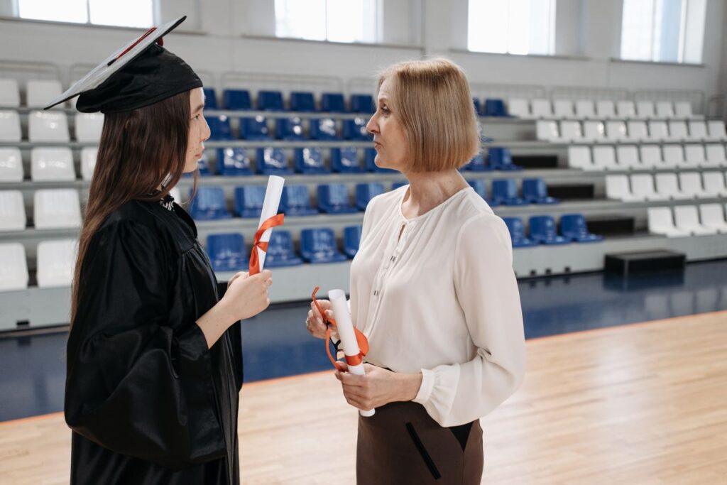 Two women discussing during a graduation ceremony, each holding diplomas.